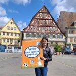 Eine lächelnde Frau mit dunklem Haar steht mit einem orangefarbenen Wir-und-Sportplätze-Plakat auf dem Esslinger Marktplatz.